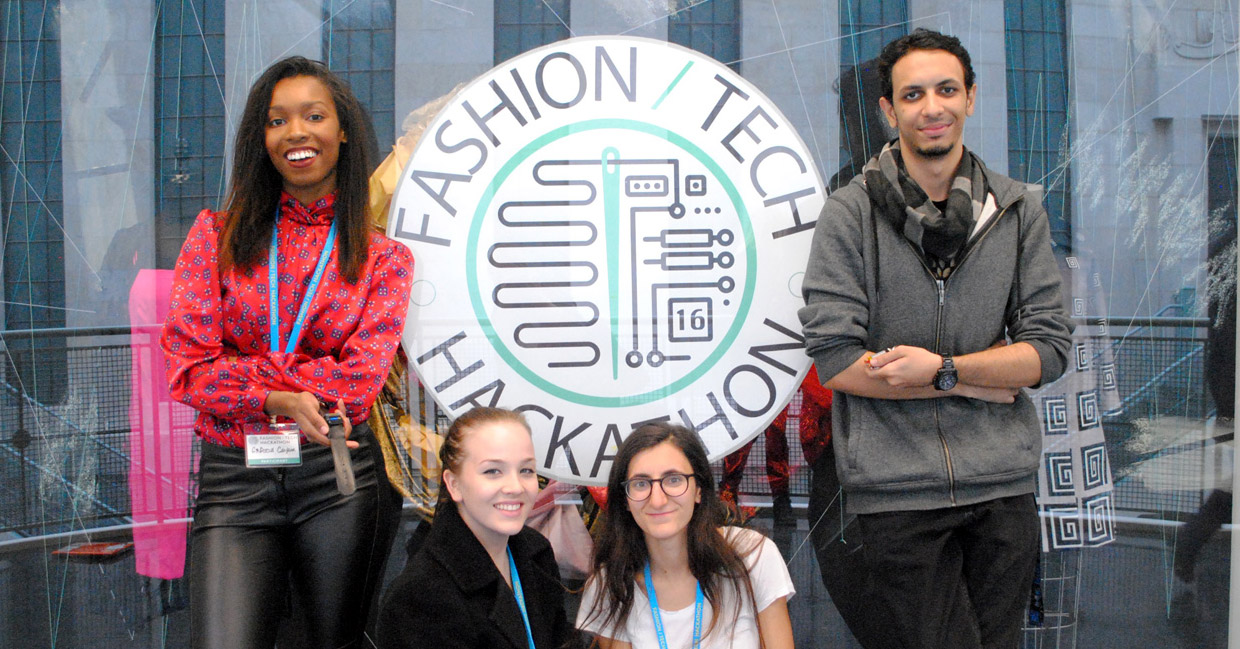 Four people posing in front of the Fashion/Tech Hackathon logo
